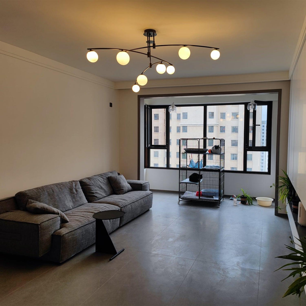 This Clérachic black chandelier glowing a warmer, yellower white in a grey living room with a sofa and a tall window.