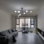 Black Clérachic chandelier lit in a cooler white tone, over a charcoal sofa in a room with a large window.