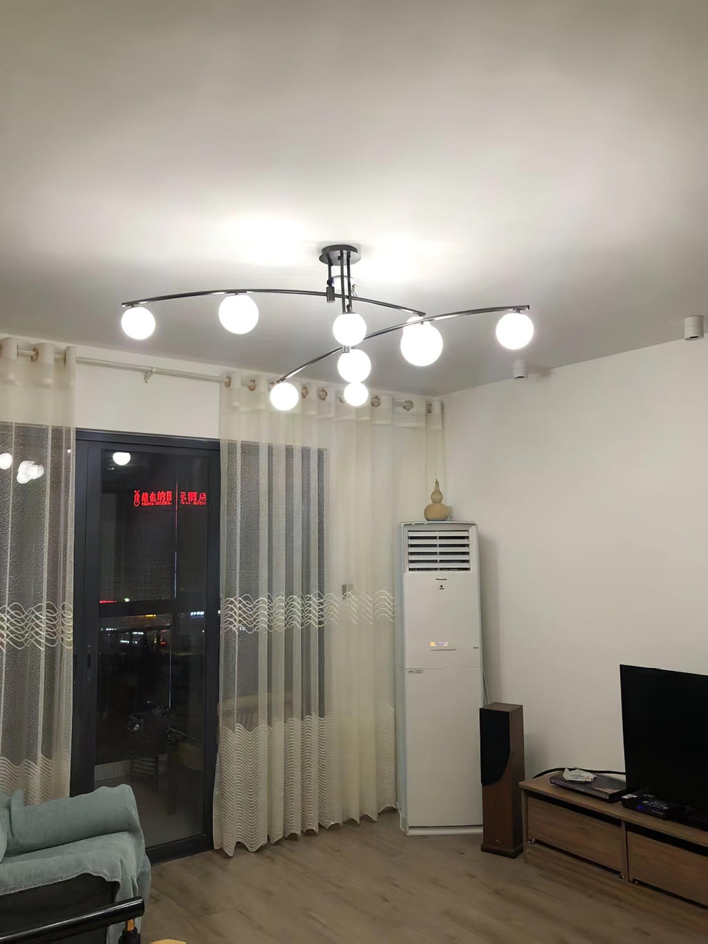 A black Clérachic chandelier lit in a plain room with sheer curtains, a wooden floor and a night view through the window.