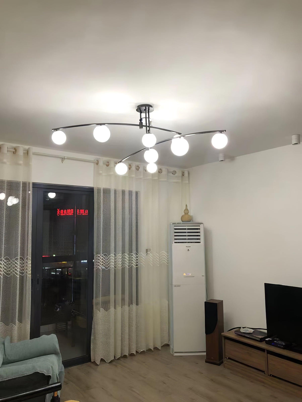 A black Clérachic chandelier lit in a plain room with sheer curtains, a wooden floor and a night view through the window.