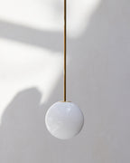 Close view of an opal sphere on a slim brass rod against a pale wall, with soft shadows falling behind this Clérachic pendant.