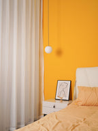 Smaller opal globe hung beside cream curtains and a yellow wall, with a bedside table and peach bedding below, a Clérachic pendant.