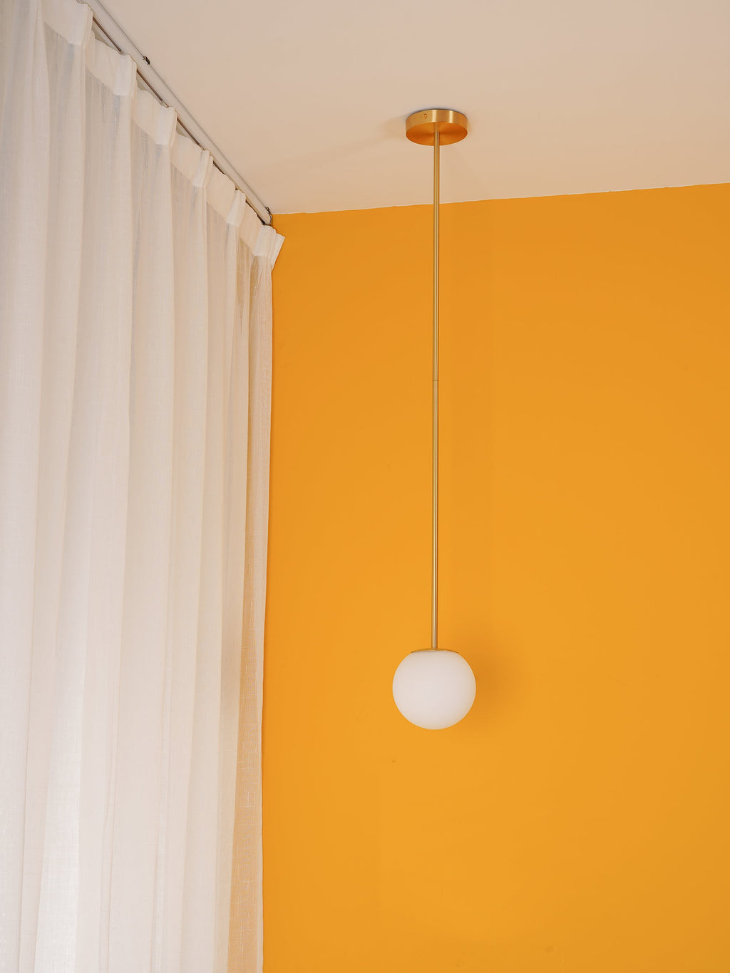 Unlit opal globe on a thin brass rod from a ceiling plate, hung in a yellow corner beside white curtains, a Clérachic pendant.