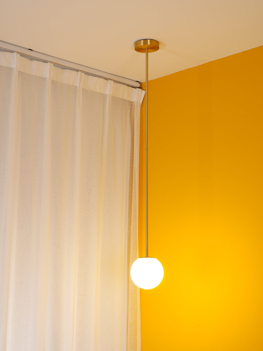 Lit opal globe on a brass rod in a yellow room corner, a Clérachic pendant glowing in front of white curtains.