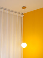 Lit opal globe on a brass rod in a yellow room corner, a Clérachic pendant glowing in front of white curtains.