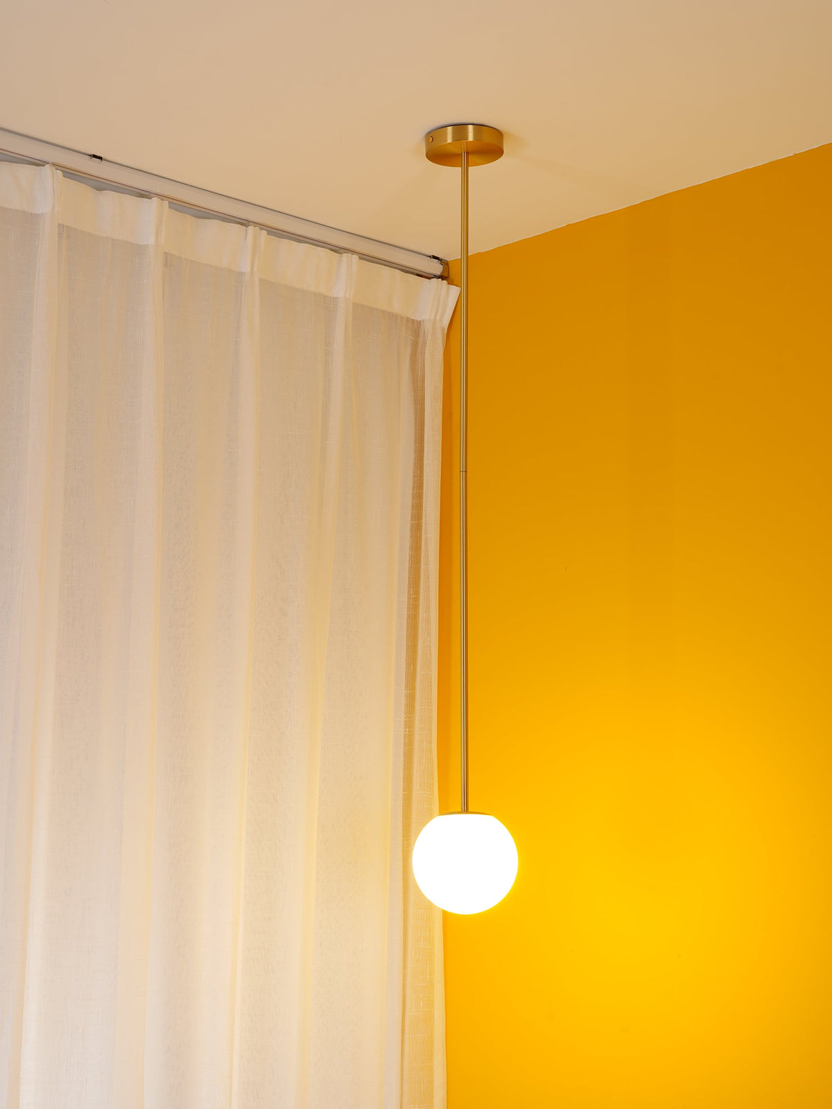Lit opal globe on a brass rod in a yellow room corner, a Clérachic pendant glowing in front of white curtains.