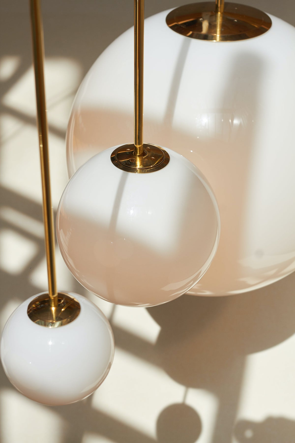 Looking up at three opal glass spheres with brass collars and rods, with sunlight and shadows on the wall behind, Clérachic pendants.