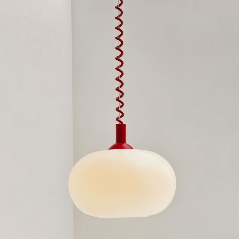 Close view of a Clérachic opal glass pendant lit with a warm cream glow, on a red coiled spring cable and cap.