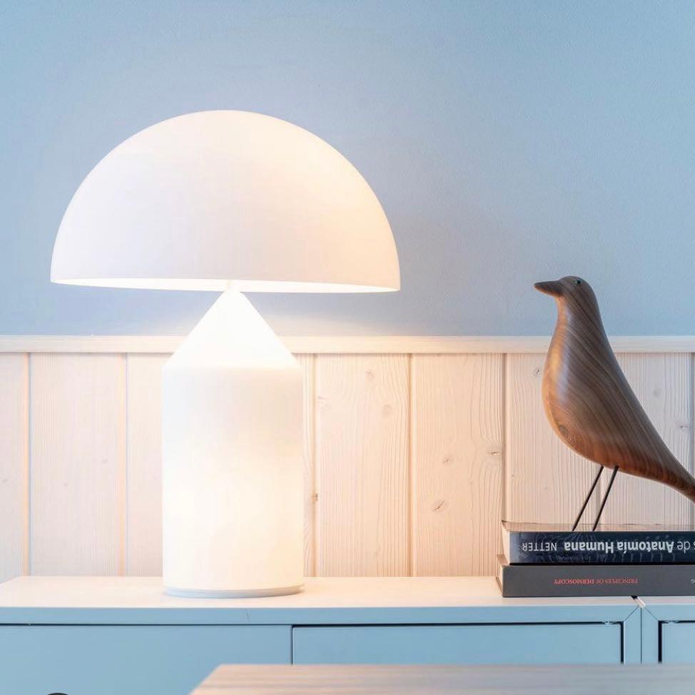A Clérachic white glass table lamp on a white sideboard against pale wood panelling, with a carved wooden bird and stacked books beside it.