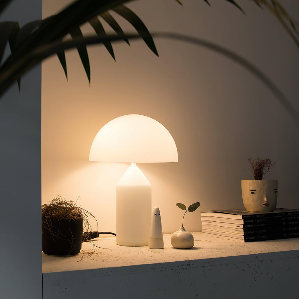 A lit Clérachic white glass table lamp on a pale concrete shelf, with small ceramic objects, a plant and stacked books.