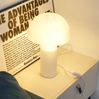The glass dome and cylinder of this Clérachic table lamp glow softly on a white bedside unit, a poster lettered in black behind.