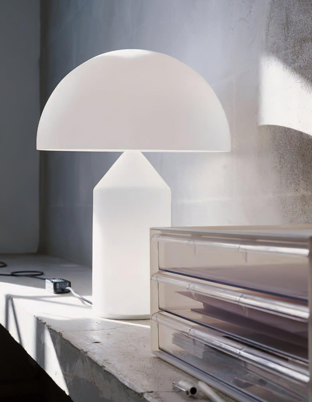 Side-lit by window light, a Clérachic white glass table lamp stands on a concrete ledge beside a clear acrylic drawer unit.