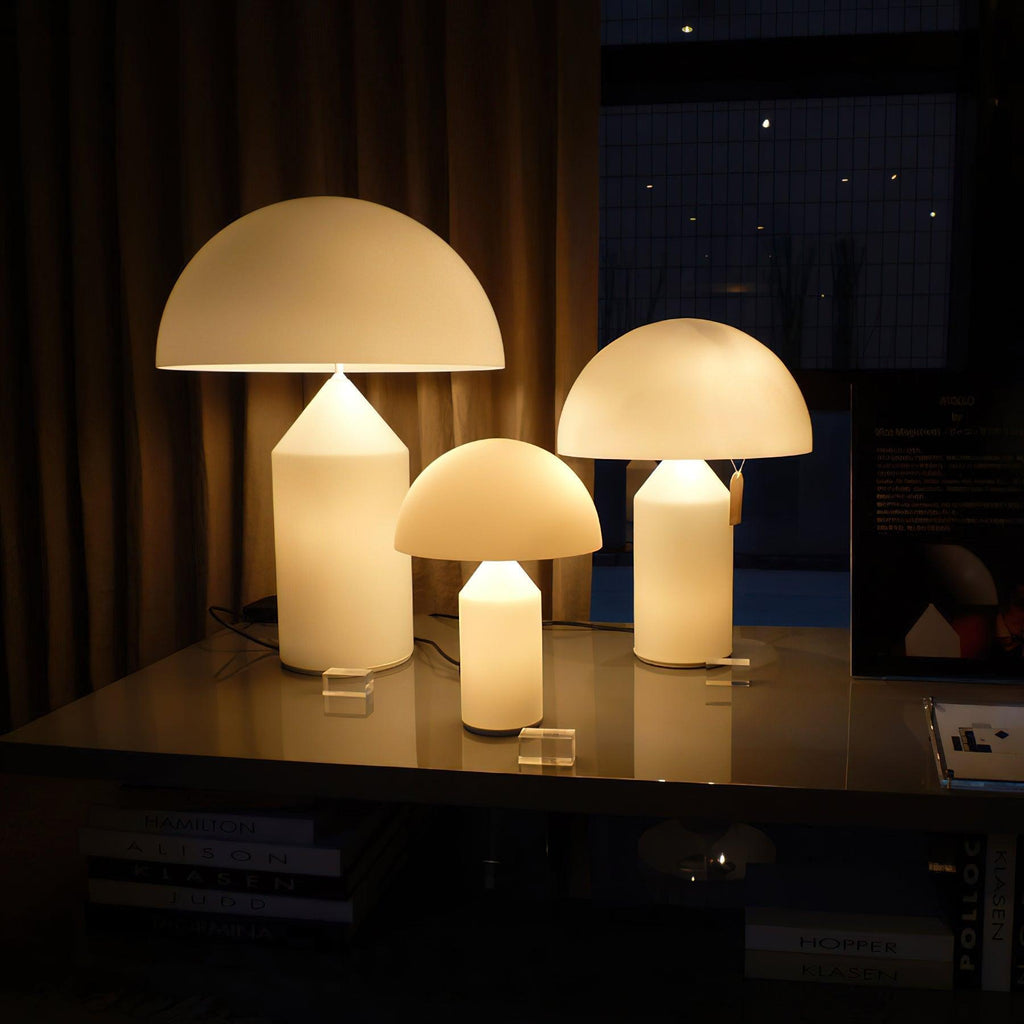 Three Clérachic opal glass table lamps in graduated sizes glow together on a dark glass shelf in front of a curtain.
