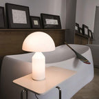A lit Clérachic opal glass table lamp on a small white side table, with a curved white sofa and a wooden paddle behind.