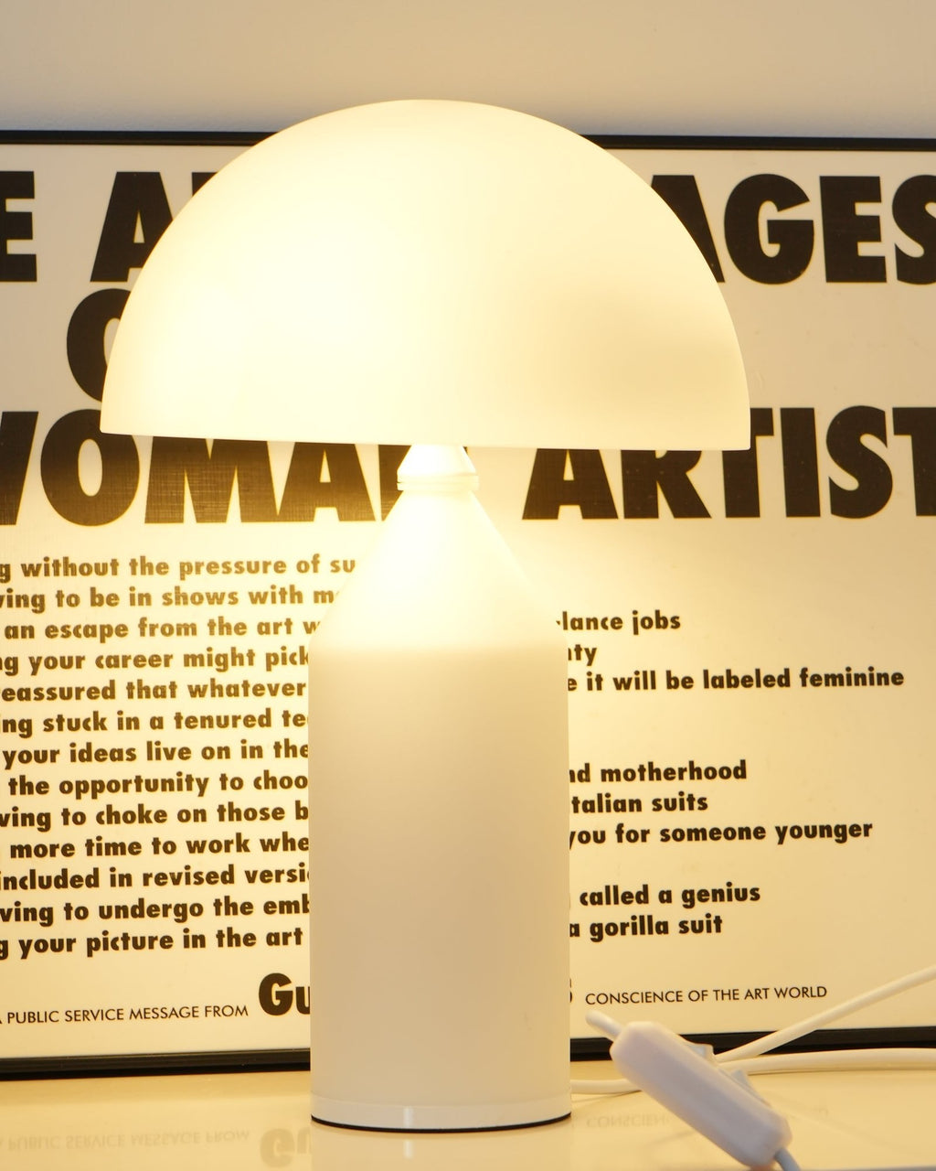 Close view of a lit Clérachic opal glass table lamp, its dome and cylinder glowing in front of a bold lettered poster.