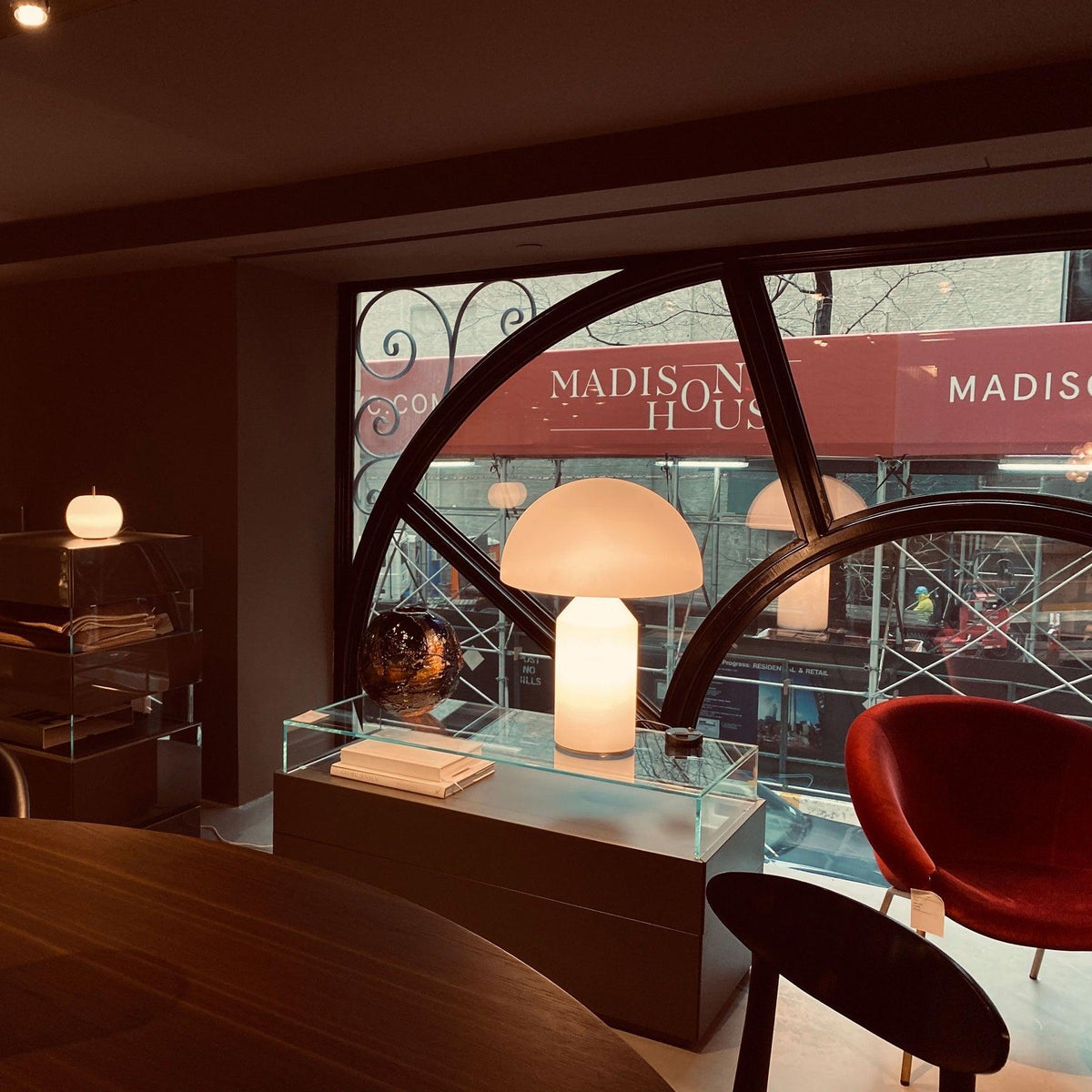 A Clérachic opal glass table lamp glows on a glass display plinth in a dim room, an arched window and red chair beyond.