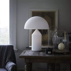 A tall Clérachic white glass table lamp on a weathered wooden table beside glass cloches, a framed print behind it.