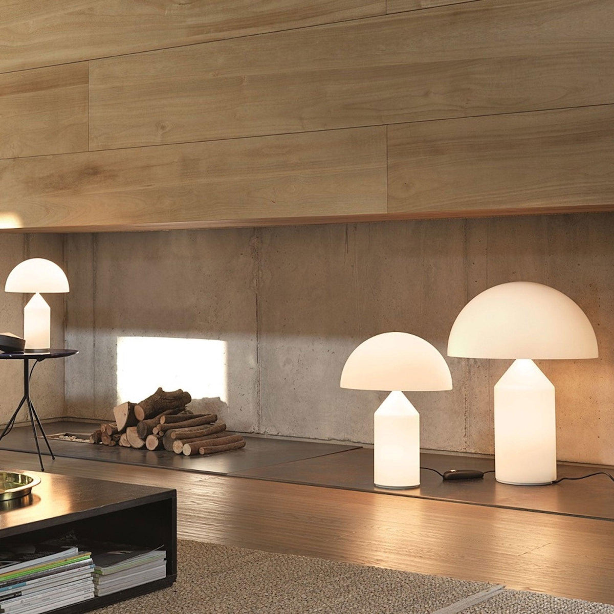 Two Clérachic opal glass table lamps of different heights glow on a low ledge in a concrete-walled room with a log store.