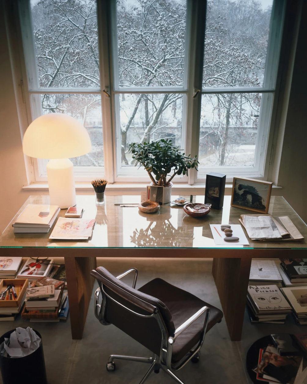 By a snow-covered window, a Clérachic opal glass table lamp stands on a glass-topped desk with books, a plant and a leather chair.