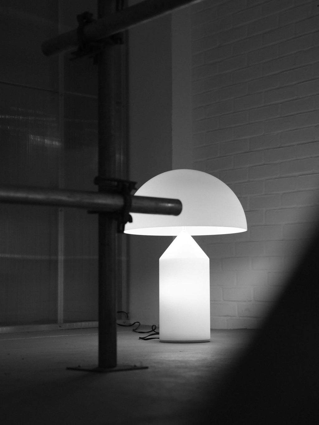 A Clérachic white glass table lamp lit in a stark black-and-white scene, with a steel pipe crossing the frame and a brick wall.