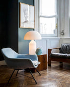 Beside a tall window in a blue-green room, this Clérachic opal glass table lamp stands on a wooden table by a pale blue armchair.
