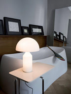 A lit Clérachic opal glass table lamp on a small white side table, a white curved sofa and a wooden paddle behind it.