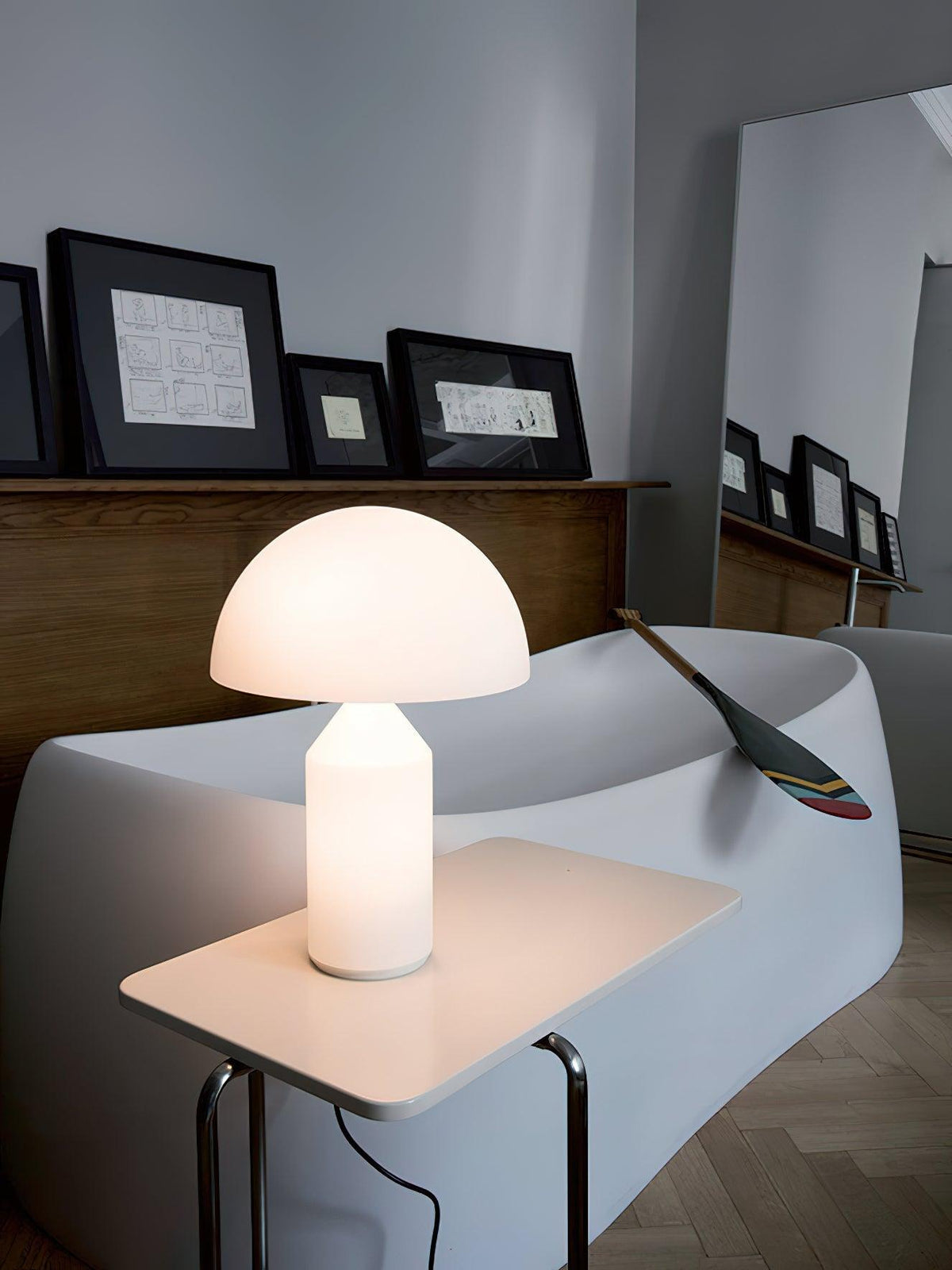 A lit Clérachic opal glass table lamp on a small white side table, a white curved sofa and a wooden paddle behind it.