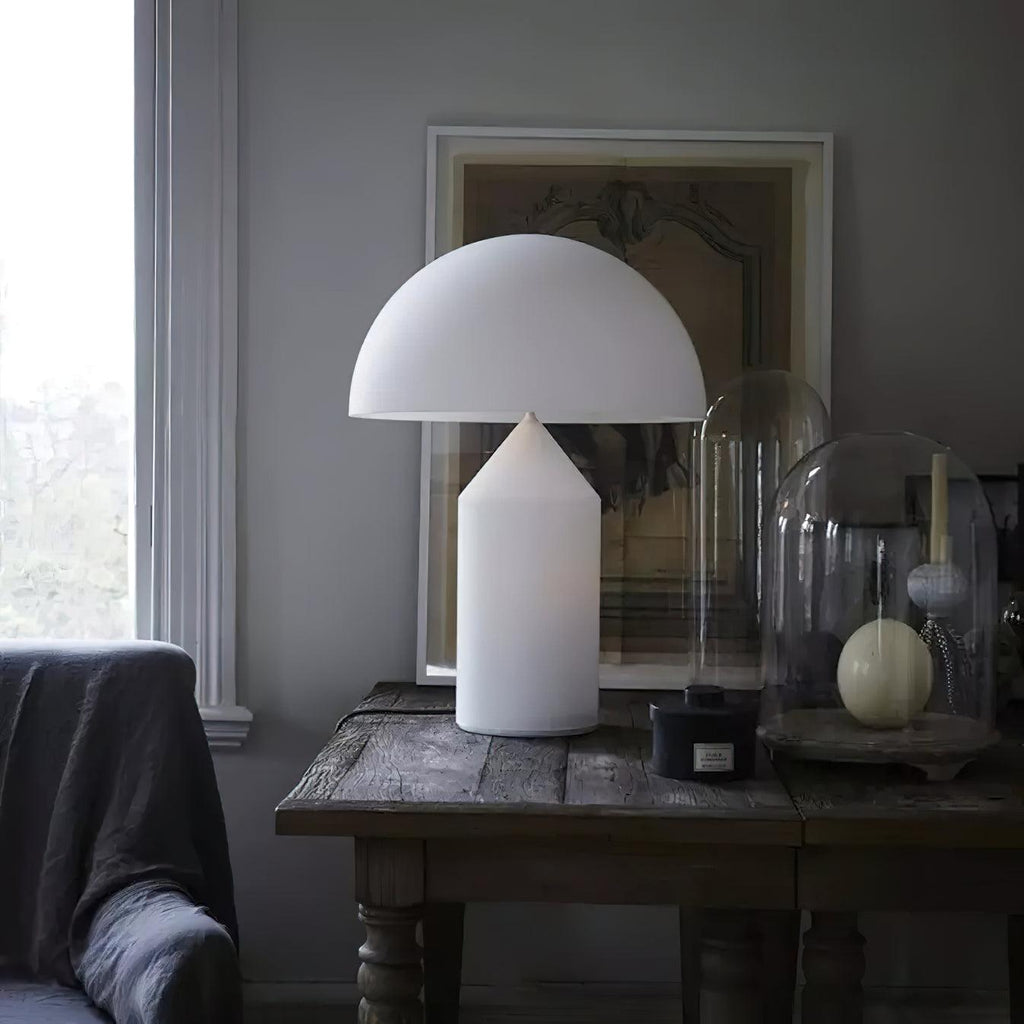 A Clérachic white glass table lamp on a weathered table with glass domes, in a grey room with a tall window.
