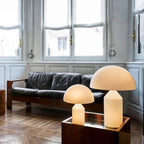 Two Clérachic opal glass table lamps glow in front of a black leather sofa, one standing on a mirrored wooden stand.