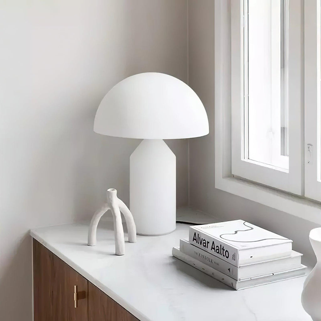 On a white counter by a window, this Clérachic white glass table lamp sits beside a sculptural white branch and an art book.