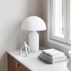 On a white counter by a window, this Clérachic white glass table lamp sits beside a sculptural white branch and an art book.