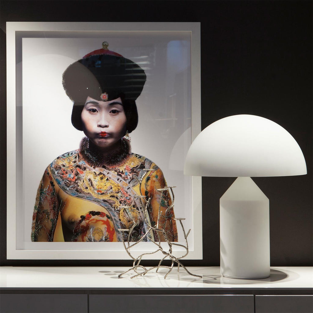 A Clérachic white glass table lamp beside a large framed portrait and a silver branch sculpture on a white sideboard.