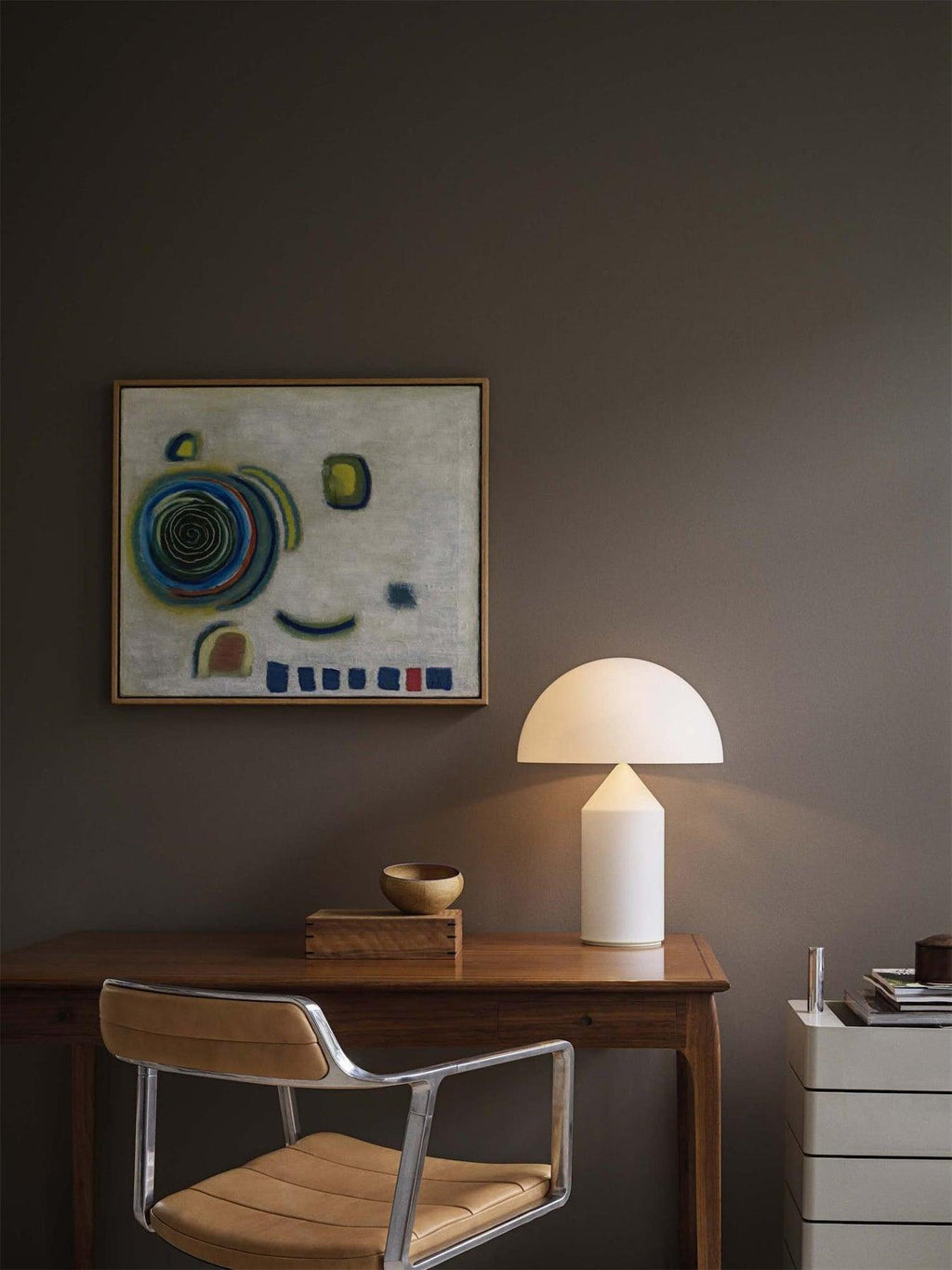 Standing by a wooden desk, this Clérachic white glass table lamp sits near a small bowl, a framed painting and a leather chair.