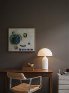 Standing by a wooden desk, this Clérachic white glass table lamp sits near a small bowl, a framed painting and a leather chair.