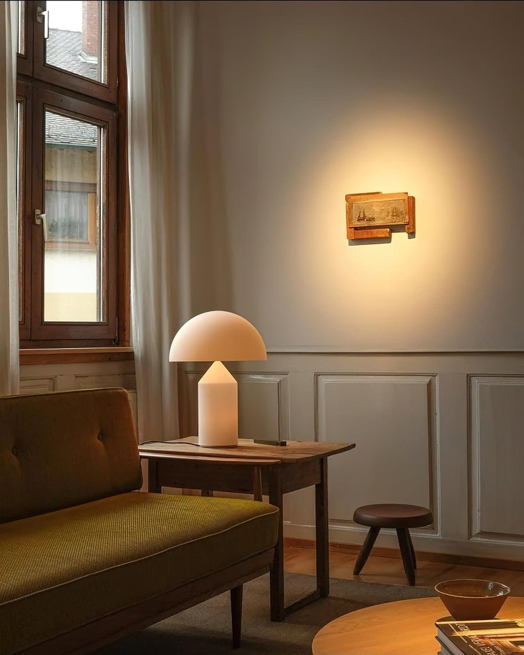 In a quiet corner with an olive-green sofa, a Clérachic white glass table lamp glows on a wooden side table beside a stool.