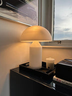 Light from the window warms the glass dome of this Clérachic table lamp, set on a black tray with a candle on a dark cabinet.