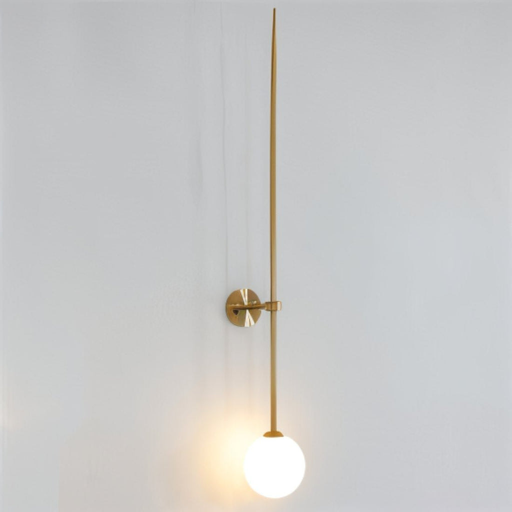 Clérachic gold wall light with a long tapered rod and a glowing white opal globe on a pale grey wall.