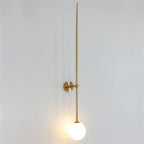 Clérachic gold wall light with a long tapered rod and a glowing white opal globe on a pale grey wall.