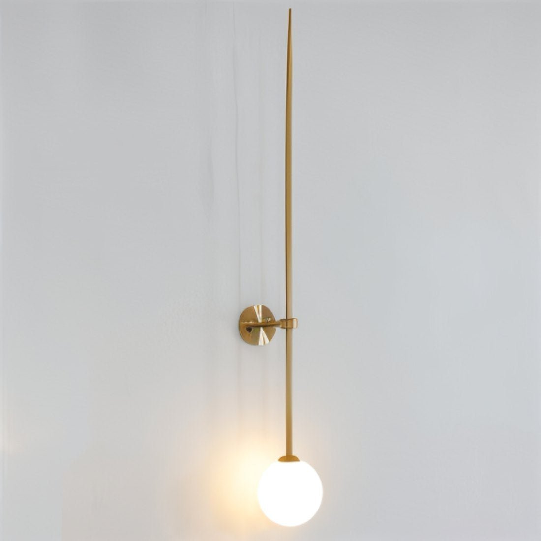 Clérachic gold wall light with a long tapered rod and a glowing white opal globe on a pale grey wall.
