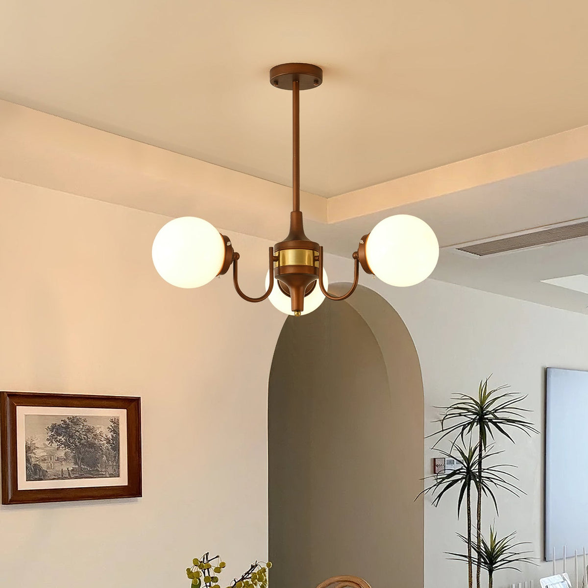 Three-globe Clérachic chandelier lit in a dining room with an arched doorway, a framed landscape print and a tall palm.
