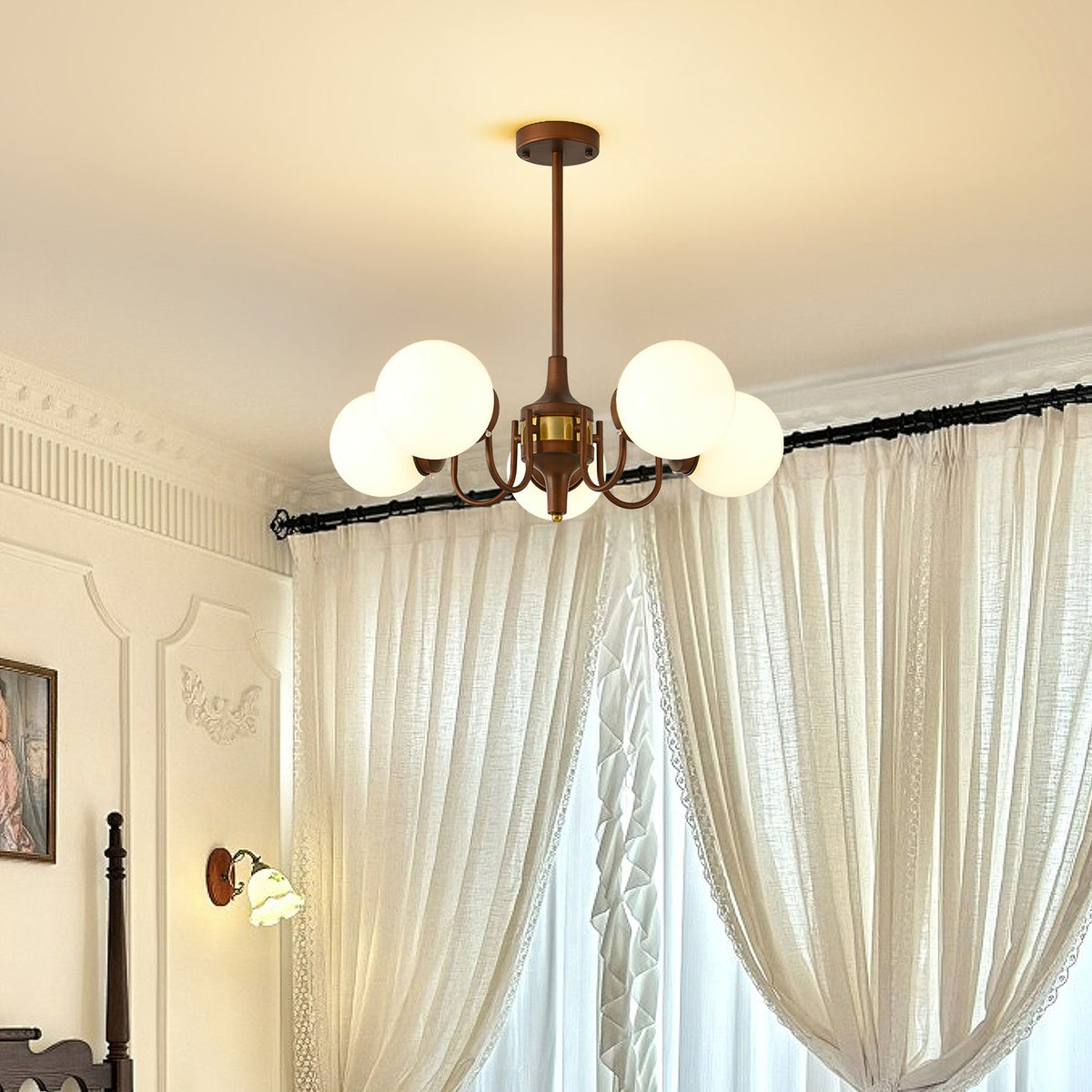 A Clérachic chandelier with white globes in front of sheer curtains and a cornice, with a small wall light and a wooden bedpost beyond.