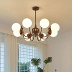 Eight-globe Clérachic chandelier lit below a white ceiling, with a palm and a gridded window behind it.