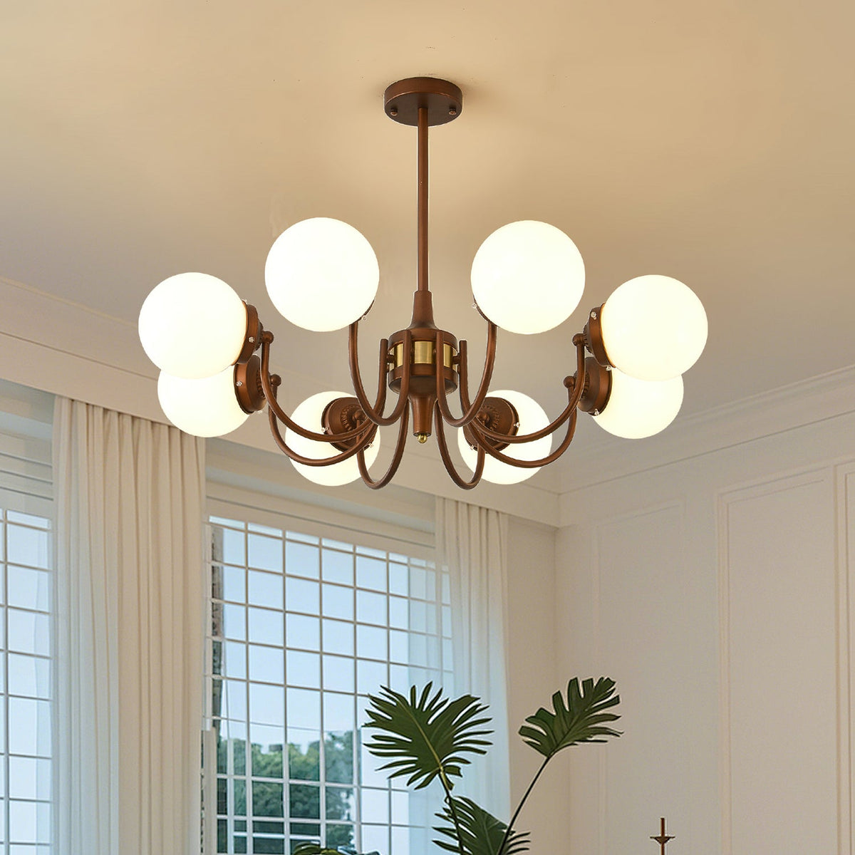 Eight-globe Clérachic chandelier lit below a white ceiling, with a palm and a gridded window behind it.