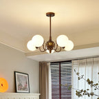 Five-globe Clérachic chandelier lit above a window with blinds and sheer curtains, with a framed floral print nearby.