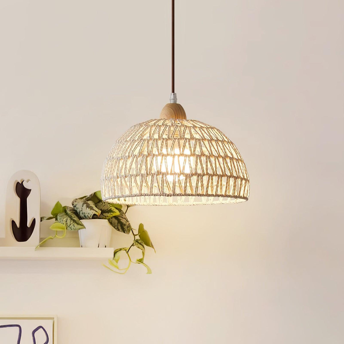 Lit Clérachic open-weave dome pendant against a cream wall, with a trailing green plant on a white shelf and a small black figure beside it.