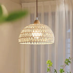 Clérachic woven dome pendant glowing warm in front of sheer white curtains, with leafy green stems in the lower corner.