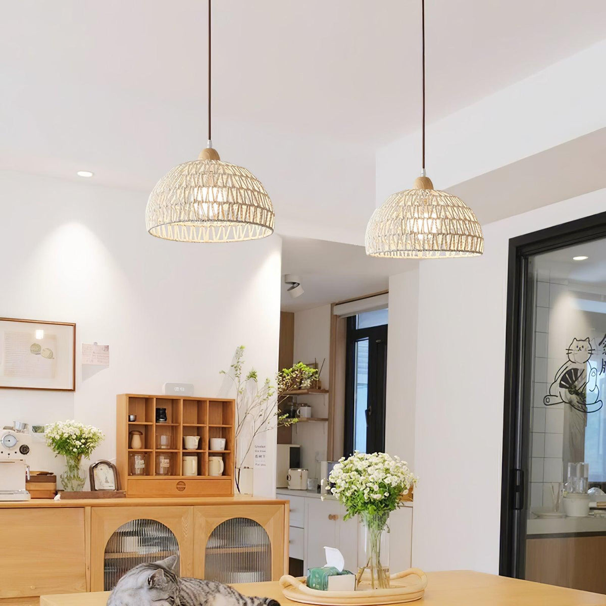 Two Clérachic woven pendants at different heights above a wooden dining area, with white flowers and a sideboard with glazed doors.