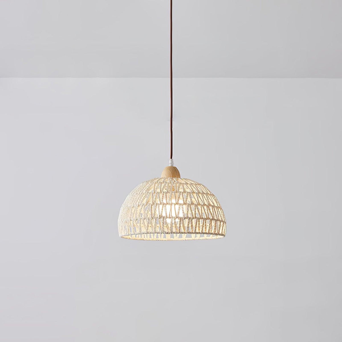 Smaller Clérachic dome pendant hanging in a plain grey room, the open weave showing a soft light inside and a brown cord above.