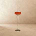 A studio view of the Clérachic floor lamp in orange glass, lit softly on a slim chrome stem and round black base.