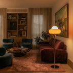 A Clérachic floor lamp in orange glass glows beside a burgundy velvet sofa, teal armchairs and a wall of bookshelves.